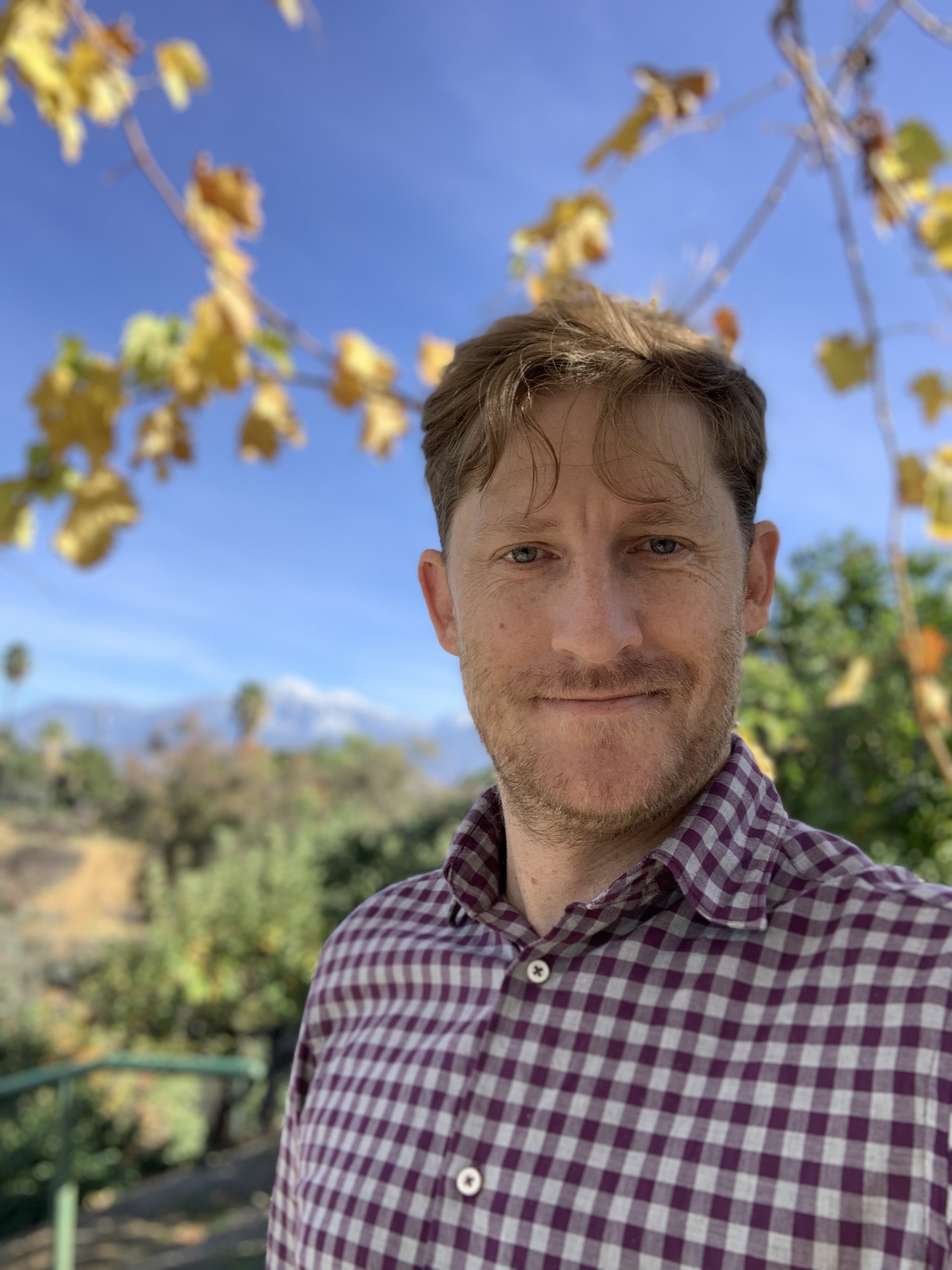 Portrait of Chris R. Becker smiling in a checked shirt, with yellow leaves and blue sky behind